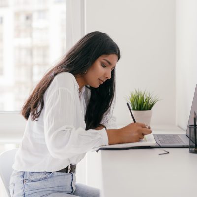 Moça na frente do computador, escreve no caderno